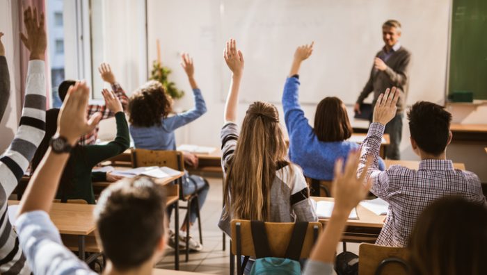 Back view of high school students raising hands on a class. Rear view of large group of students raising their hands to answer the question on a class.